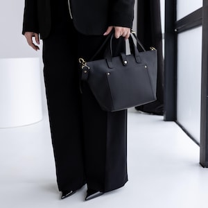 May include: A black leather tote bag with gold-colored hardware, including handles and a shoulder strap. The bag has a structured design with a rectangular shape and a top handle. The bag is held by a person wearing a black blazer and pants.