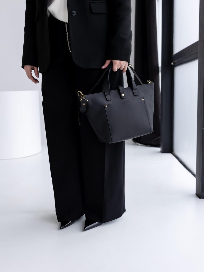 May include: A black leather tote bag with gold-colored hardware, including handles and a shoulder strap. The bag has a structured design with a rectangular shape and a top handle. The bag is held by a person wearing a black blazer and pants.