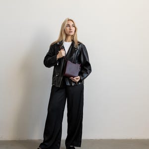 Pode incluir: Uma pessoa vestindo uma jaqueta de couro preta, camiseta branca e cal&ccedil;as pretas de perna larga. Ela est&aacute; segurando uma bolsa transversal bord&ocirc;. O traje &eacute; completado com mocassins pretos.