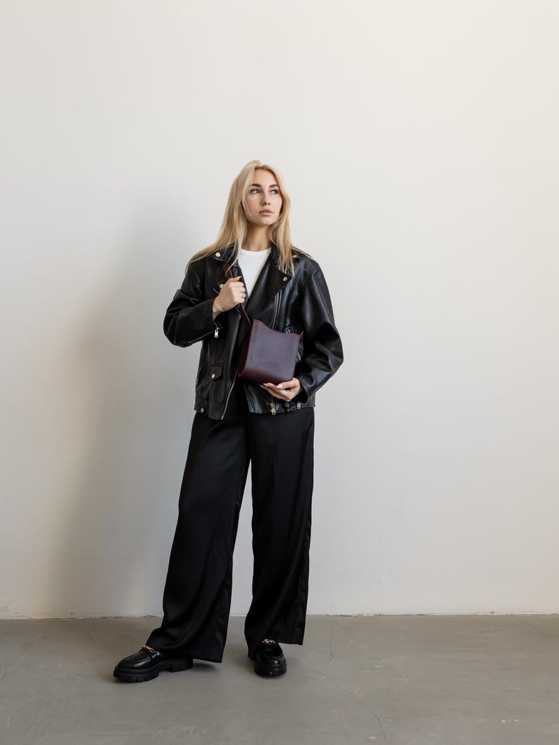 Pode incluir: Uma pessoa vestindo uma jaqueta de couro preta, camiseta branca e cal&ccedil;as pretas de perna larga. Ela est&aacute; segurando uma bolsa transversal bord&ocirc;. O traje &eacute; completado com mocassins pretos.