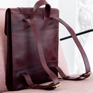 May include: A burgundy leather backpack with adjustable shoulder straps and a top handle. The bag features a rectangular shape with a flap closure and brass-colored hardware. The backpack is shown on a pink surface.