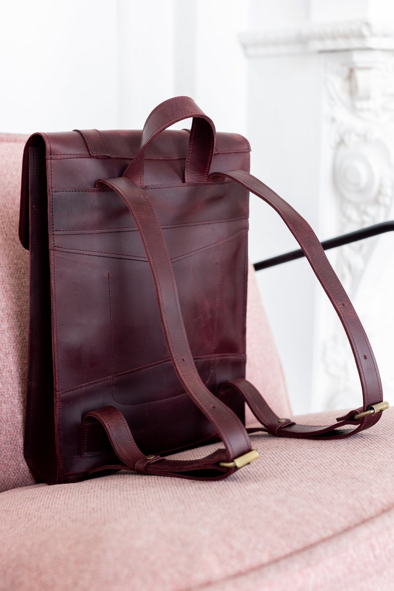 May include: A burgundy leather backpack with adjustable shoulder straps and a top handle. The bag features a rectangular shape with a flap closure and brass-colored hardware. The backpack is shown on a pink surface.