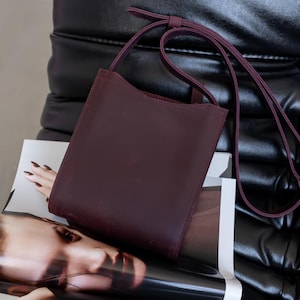 Pode incluir: Uma bolsa transversal de couro bord&ocirc; escuro com uma al&ccedil;a longa e fina. A bolsa &eacute; retangular com uma borda superior curva. Ela repousa sobre uma revista com o rosto de uma mulher na capa, contra uma cadeira de couro preta.