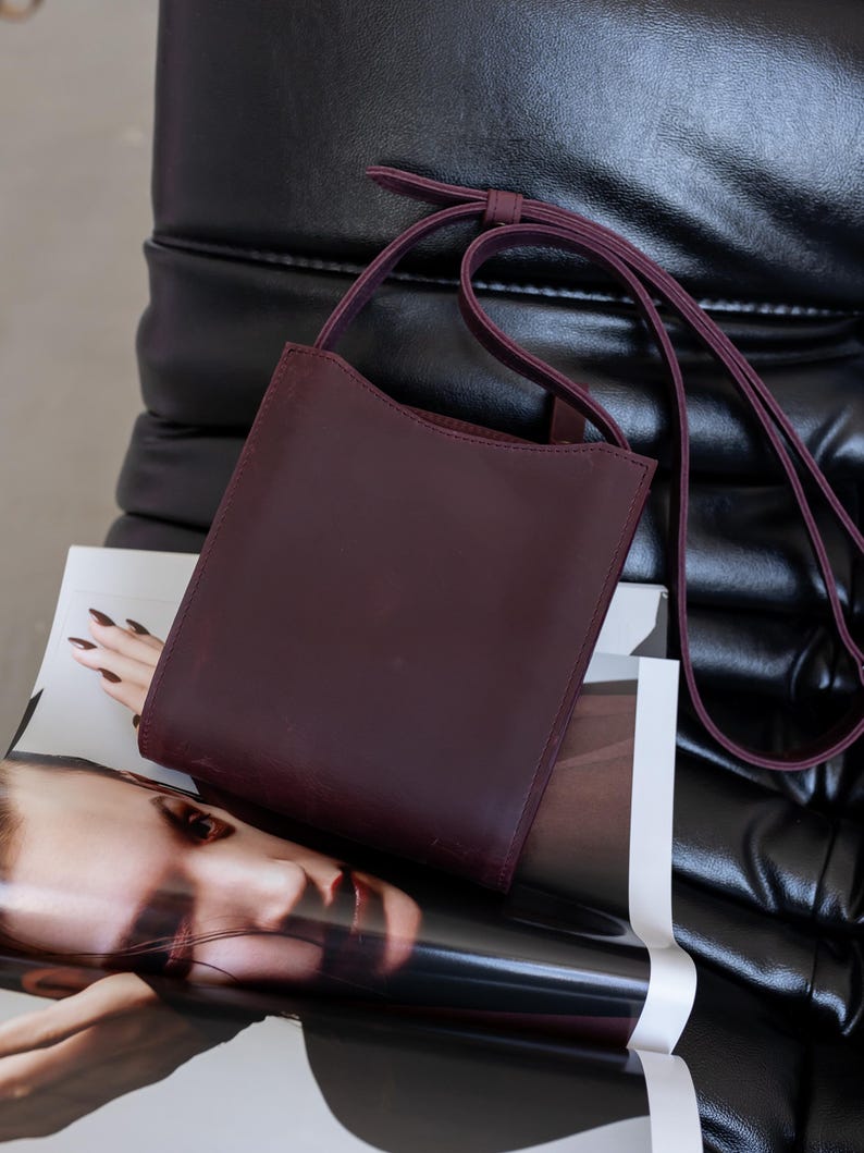 Pode incluir: Uma bolsa transversal de couro bord&ocirc; escuro com uma al&ccedil;a longa e fina. A bolsa &eacute; retangular com uma borda superior curva. Ela repousa sobre uma revista com o rosto de uma mulher na capa, contra uma cadeira de couro preta.