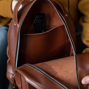 Peut inclure: Sac &agrave; dos en cuir marron ouvert, avec une fermeture &eacute;clair argent&eacute;e et une poign&eacute;e sup&eacute;rieure. L'int&eacute;rieur r&eacute;v&egrave;le une poche et une paire de lunettes de soleil noires. Le sac a une forme arrondie et une petite poche lat&eacute;rale.