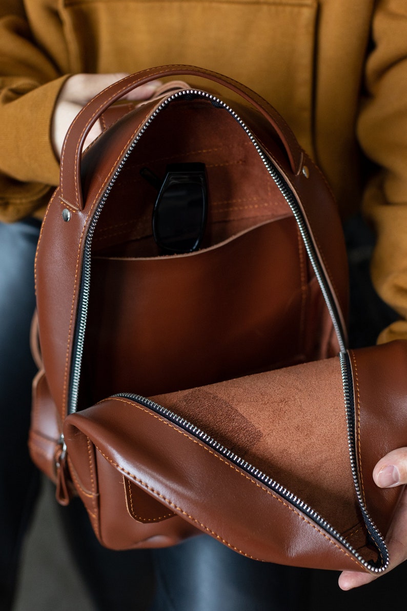 Peut inclure: Sac &agrave; dos en cuir marron ouvert, avec une fermeture &eacute;clair argent&eacute;e et une poign&eacute;e sup&eacute;rieure. L'int&eacute;rieur r&eacute;v&egrave;le une poche et une paire de lunettes de soleil noires. Le sac a une forme arrondie et une petite poche lat&eacute;rale.