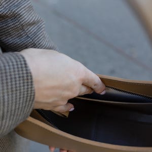Puede incluir: Un bolso de mano de cuero color canela con cremallera e interior negros. El bolso est&aacute; abierto, revelando el interior. La mano de una persona sostiene el bolso abierto. La persona lleva una blazer a cuadros marr&oacute;n y gris.