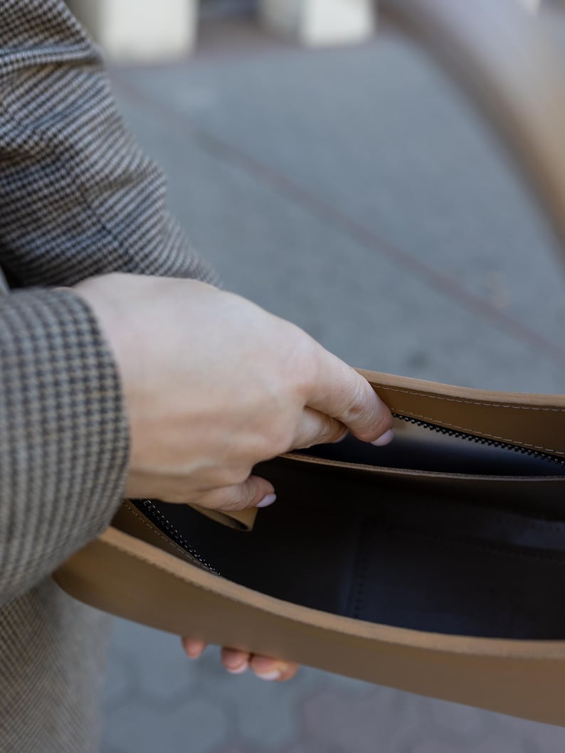Puede incluir: Un bolso de mano de cuero color canela con cremallera e interior negros. El bolso est&aacute; abierto, revelando el interior. La mano de una persona sostiene el bolso abierto. La persona lleva una blazer a cuadros marr&oacute;n y gris.