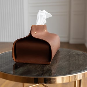 May include: A brown leather tissue box with a curved design and brass rivets. White tissues are visible at the top. The tissue holder is on a round table with a dark marble top and gold-colored metal legs. Home decor.