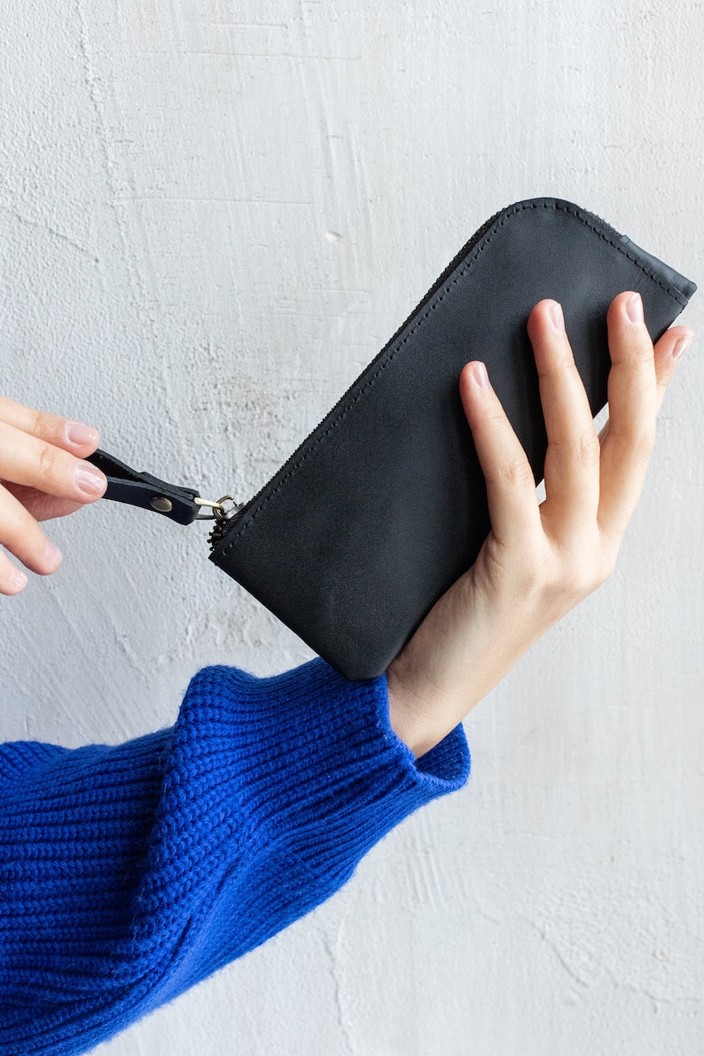 May include: A black leather pencil case with a zipper closure and a wrist strap. The case is held against a white textured wall. The person holding the case is wearing a blue sweater.