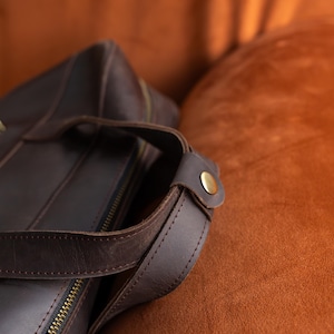 May include: A dark brown leather bag featuring a top handle secured with a metallic brass-colored snap button. The bag includes a visible zipper closure along the top edge, with visible stitching along the edges of the leather handle and body.