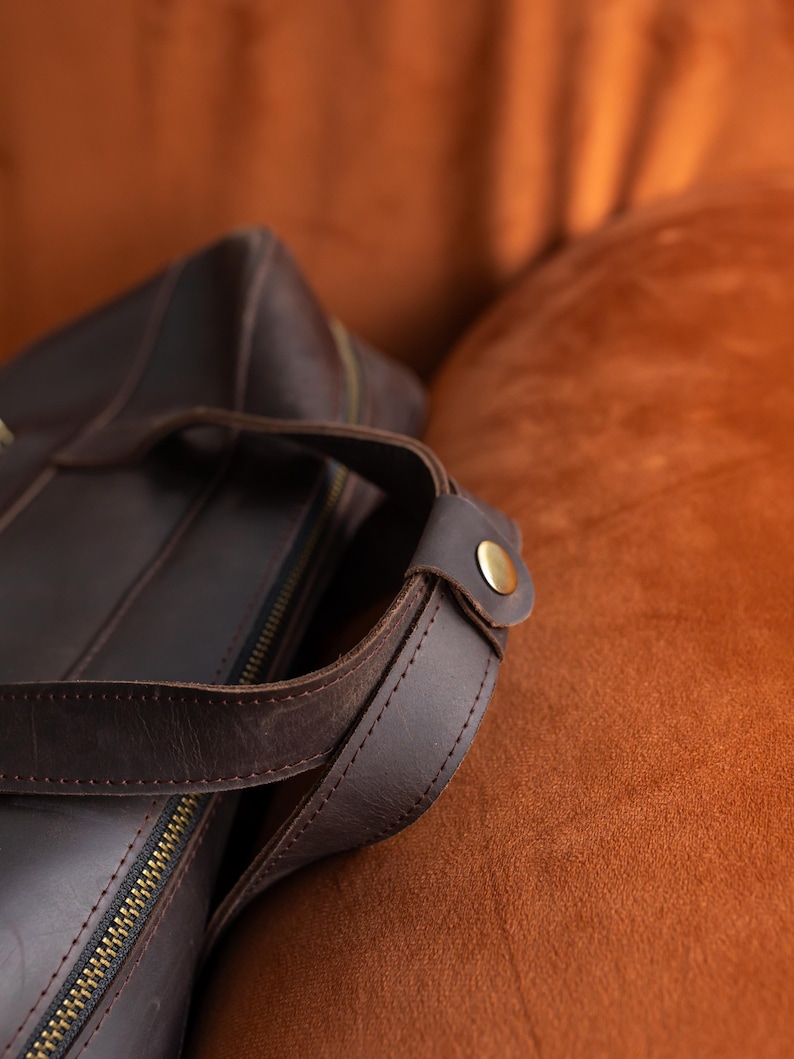 May include: A dark brown leather bag featuring a top handle secured with a metallic brass-colored snap button. The bag includes a visible zipper closure along the top edge, with visible stitching along the edges of the leather handle and body.