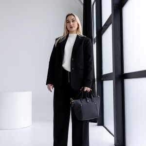May include: A person wearing a black blazer, black pants, and a white sweater, holding a black handbag. The outfit is set against a white background with a large window and black trim.