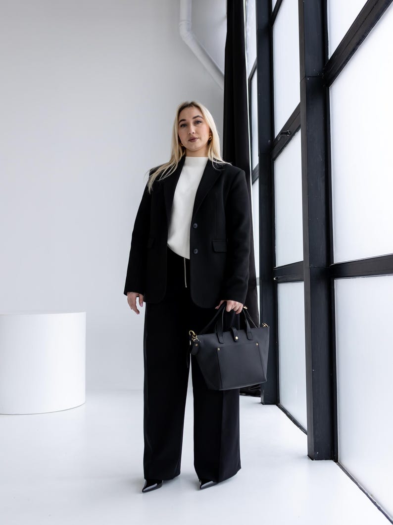 May include: A person wearing a black blazer, black pants, and a white sweater, holding a black handbag. The outfit is set against a white background with a large window and black trim.
