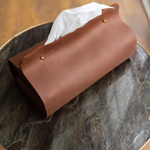May include: A brown leather tissue box cover with a curved design. The cover is secured with brass-colored rivets and holds white tissues. The tissue box is on a round table with a dark gray marble top and a gold-colored metal frame.
