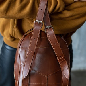 Peut inclure: Un sac &agrave; dos en cuir marron avec des bretelles r&eacute;glables et des boucles argent&eacute;es. Le sac pr&eacute;sente un motif g&eacute;om&eacute;trique cousu et une fermeture &eacute;clair. Les bretelles sont fix&eacute;es en haut du sac et sont en cuir marron.