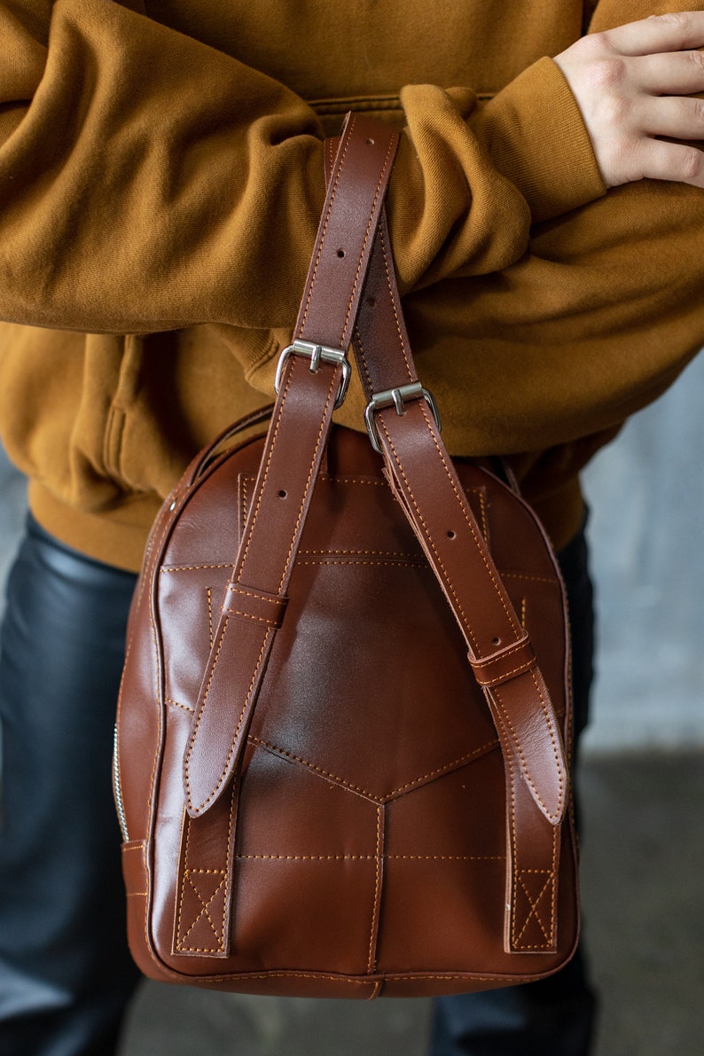 Peut inclure: Un sac &agrave; dos en cuir marron avec des bretelles r&eacute;glables et des boucles argent&eacute;es. Le sac pr&eacute;sente un motif g&eacute;om&eacute;trique cousu et une fermeture &eacute;clair. Les bretelles sont fix&eacute;es en haut du sac et sont en cuir marron.