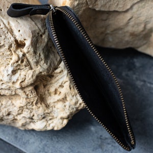 May include: Black leather zippered pouch with a brass-colored zipper. The pouch is open, revealing a dark interior. A small leather loop is attached to the zipper pull. The pouch is set against a neutral background.