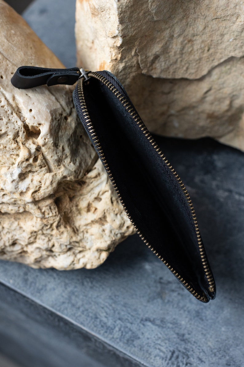 May include: Black leather zippered pouch with a brass-colored zipper. The pouch is open, revealing a dark interior. A small leather loop is attached to the zipper pull. The pouch is set against a neutral background.