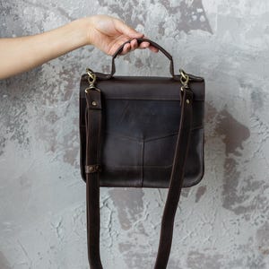 May include: A dark brown leather satchel with a top handle and adjustable shoulder strap. The bag has a rectangular shape with a flap closure and brass-colored hardware. The bag is held by a hand against a textured gray background.