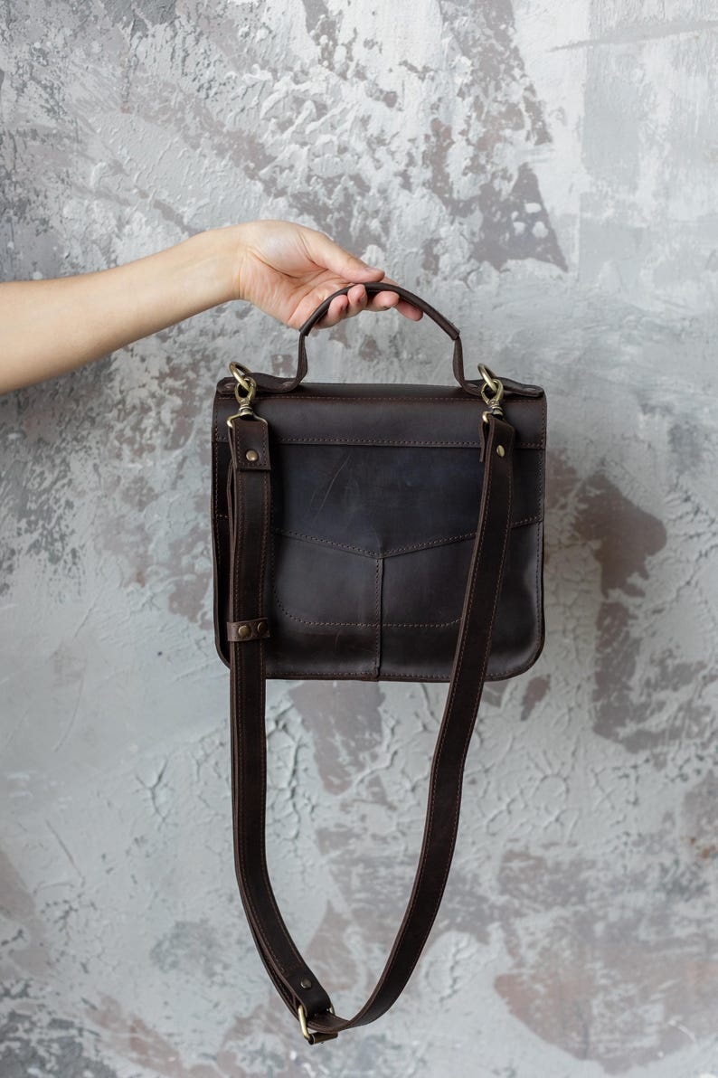 May include: A dark brown leather satchel with a top handle and adjustable shoulder strap. The bag has a rectangular shape with a flap closure and brass-colored hardware. The bag is held by a hand against a textured gray background.