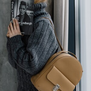 May include: A tan leather backpack with a small front pocket and silver clasp. The person is wearing a gray cable-knit turtleneck sweater and holding a book titled "Peter Lindbergh" with a black and white photograph on the cover.