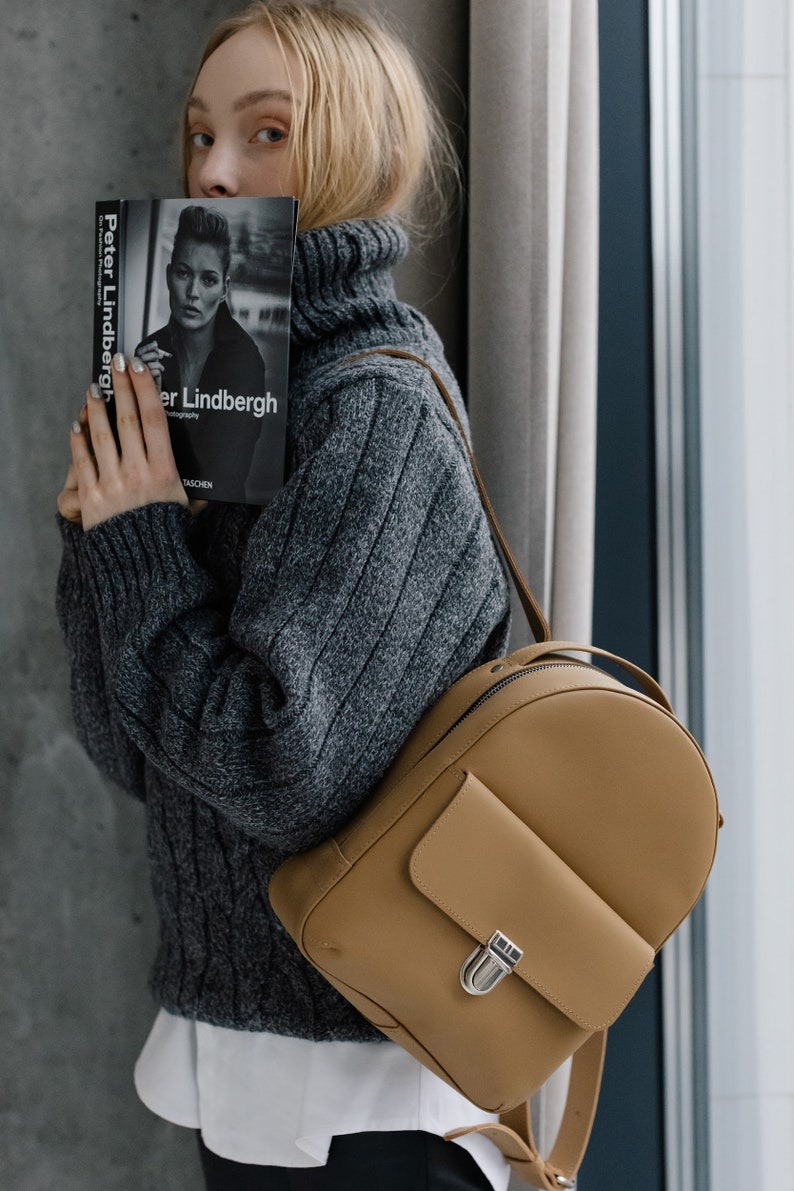 May include: A tan leather backpack with a small front pocket and silver clasp. The person is wearing a gray cable-knit turtleneck sweater and holding a book titled "Peter Lindbergh" with a black and white photograph on the cover.