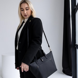 May include: A black leather tote bag with a rectangular shape and a shoulder strap. The bag has a front pocket and gold-colored hardware. The bag is being carried by a person wearing a black blazer and pants.