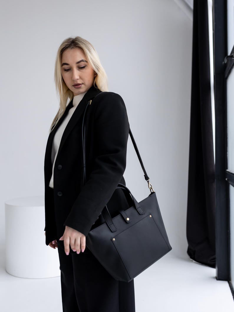 May include: A black leather tote bag with a rectangular shape and a shoulder strap. The bag has a front pocket and gold-colored hardware. The bag is being carried by a person wearing a black blazer and pants.