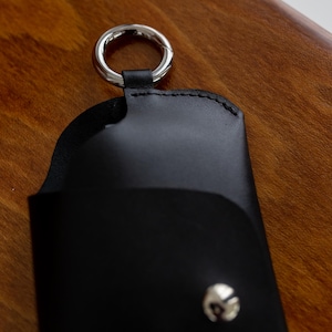 Pode incluir: Estojo de &oacute;culos em couro preto com um anel de metal prateado e fecho de press&atilde;o. O estojo foi projetado para guardar &oacute;culos e tem uma borda costurada. O fundo &eacute; uma superf&iacute;cie de madeira.