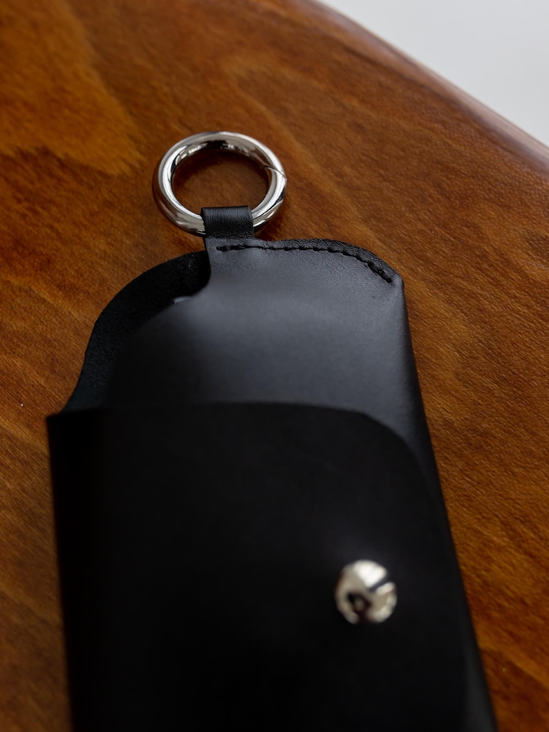 Pode incluir: Estojo de &oacute;culos em couro preto com um anel de metal prateado e fecho de press&atilde;o. O estojo foi projetado para guardar &oacute;culos e tem uma borda costurada. O fundo &eacute; uma superf&iacute;cie de madeira.
