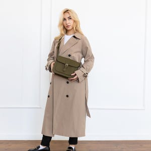 May include: An olive green crossbody bag with a buckle closure. The bag is held in front of a person wearing a tan trench coat and black pants. The coat has a double-breasted design and a belt. The person is wearing black loafers.
