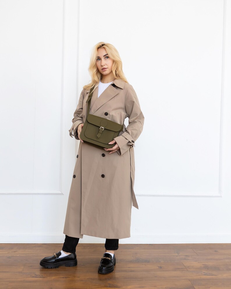 May include: An olive green crossbody bag with a buckle closure. The bag is held in front of a person wearing a tan trench coat and black pants. The coat has a double-breasted design and a belt. The person is wearing black loafers.