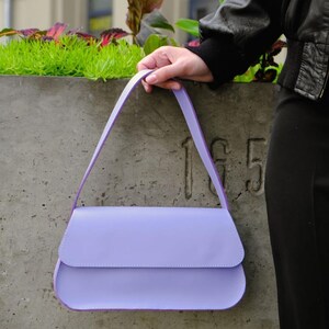 May include: A light purple shoulder bag with a smooth, solid-colored exterior, a curved flap closure, and a single matching strap. The bag features a structured, rectangular silhouette with rounded bottom corners and clean, minimalist stitching.