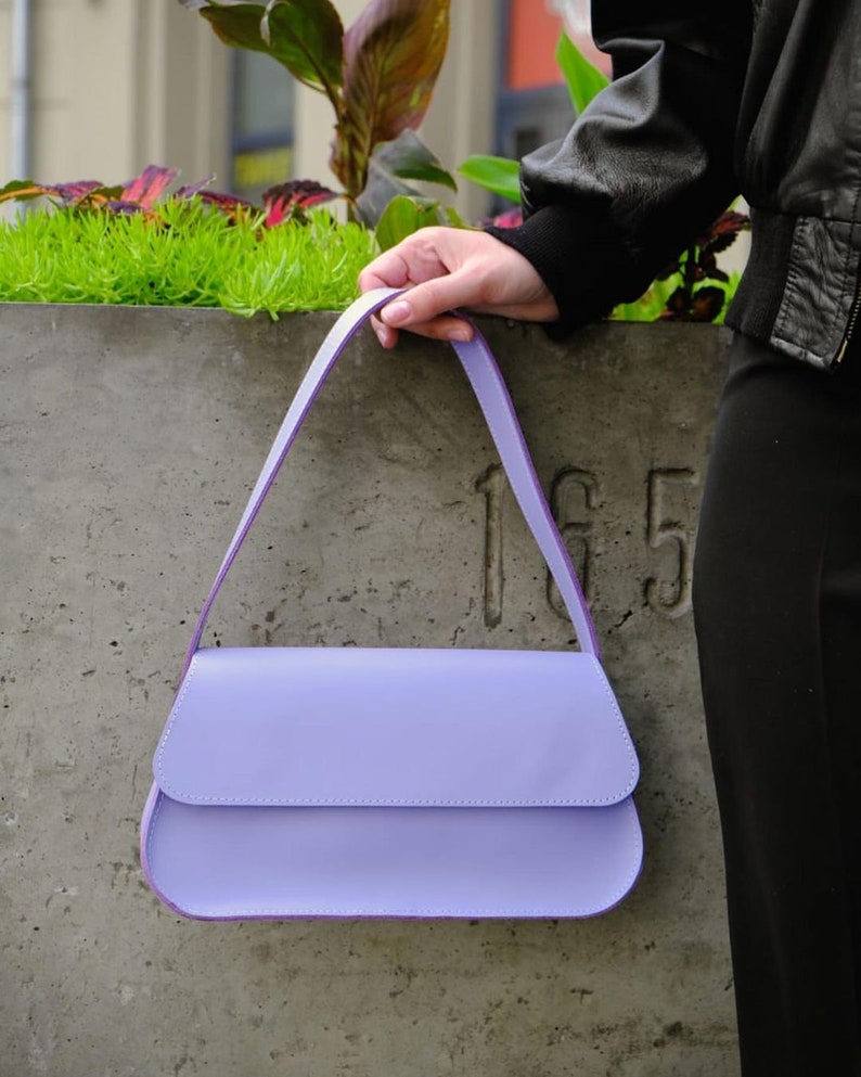 May include: A light purple shoulder bag with a smooth, solid-colored exterior, a curved flap closure, and a single matching strap. The bag features a structured, rectangular silhouette with rounded bottom corners and clean, minimalist stitching.