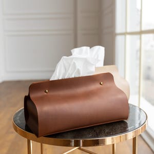 May include: A brown leather tissue box cover with gold-colored rivets and a curved design. The tissue box is on a small, round table with a gold-colored frame and a dark top. White tissues are visible.