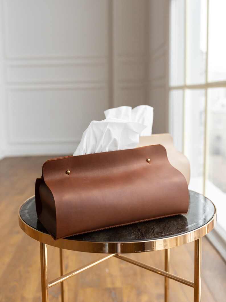 May include: A brown leather tissue box cover with gold-colored rivets and a curved design. The tissue box is on a small, round table with a gold-colored frame and a dark top. White tissues are visible.