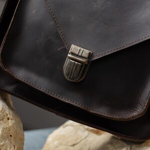 May include: A dark brown leather satchel bag with a metal clasp. The bag has a flap closure and visible stitching along the edges. The leather appears to have a distressed finish, adding to its rustic appeal.