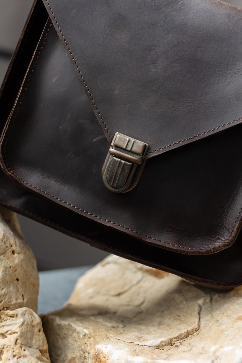 May include: A dark brown leather satchel bag with a metal clasp. The bag has a flap closure and visible stitching along the edges. The leather appears to have a distressed finish, adding to its rustic appeal.
