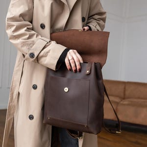 May include: A dark brown leather backpack featuring a large front pocket with a gold-toned magnetic snap closure, a fold-over flap top, and adjustable shoulder straps. The bag is constructed with visible stitching and brass-colored metal hardware.