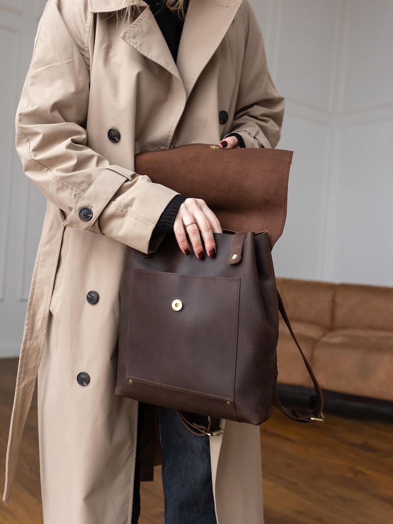 May include: A dark brown leather backpack featuring a large front pocket with a gold-toned magnetic snap closure, a fold-over flap top, and adjustable shoulder straps. The bag is constructed with visible stitching and brass-colored metal hardware.