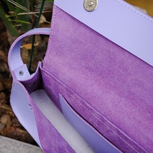 May include: A lavender leather shoulder bag with a smooth exterior and a textured, suede-like interior. The bag features a flap closure secured by a silver-toned magnetic snap button and includes an internal pocket with visible white stitching.