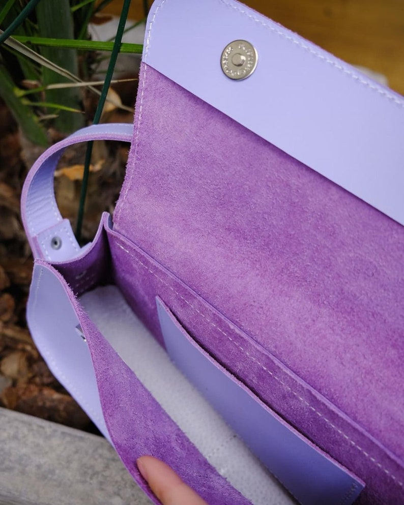 May include: A lavender leather shoulder bag with a smooth exterior and a textured, suede-like interior. The bag features a flap closure secured by a silver-toned magnetic snap button and includes an internal pocket with visible white stitching.