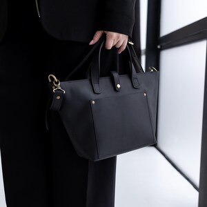 May include: A black handbag with gold-colored hardware is held by a person. The bag has a rectangular shape with a small front pocket and two handles. The bag also has a detachable shoulder strap.