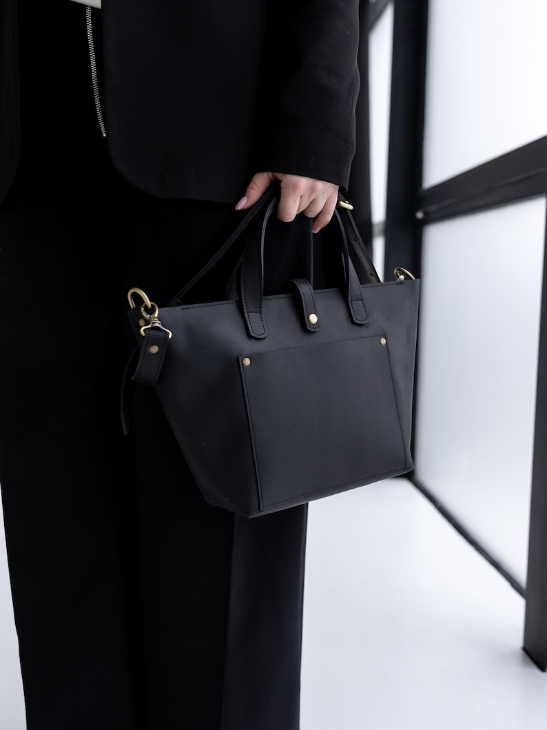 May include: A black handbag with gold-colored hardware is held by a person. The bag has a rectangular shape with a small front pocket and two handles. The bag also has a detachable shoulder strap.
