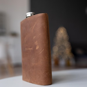 Könnte beinhalten: Eine braune Lederflasche mit silbernem Verschluss. Die Flasche hat eine rechteckige Form mit abgerundeten Kanten und einer strukturierten Oberfläche. Der Hintergrund ist verschwommen, mit einem Hauch von Weihnachtsbaum.