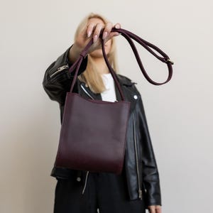 Pode incluir: Uma bolsa transversal de couro bord&ocirc; com formato retangular e uma al&ccedil;a longa e fina. A bolsa &eacute; segurada por uma pessoa, e o fundo &eacute; de cor neutra.