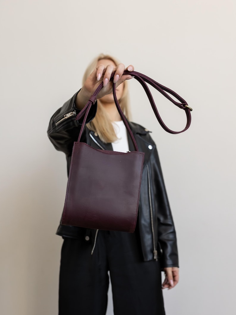 Pode incluir: Uma bolsa transversal de couro bord&ocirc; com formato retangular e uma al&ccedil;a longa e fina. A bolsa &eacute; segurada por uma pessoa, e o fundo &eacute; de cor neutra.