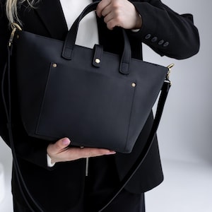 May include: A black leather tote bag with a shoulder strap and top handles. The bag has a front pocket and gold-colored hardware. The bag is held by a person wearing a black blazer and a white shirt.
