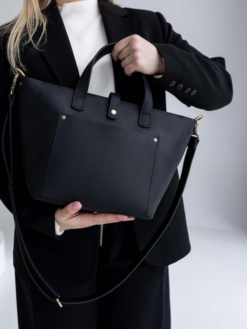 May include: A black leather tote bag with a shoulder strap and top handles. The bag has a front pocket and gold-colored hardware. The bag is held by a person wearing a black blazer and a white shirt.